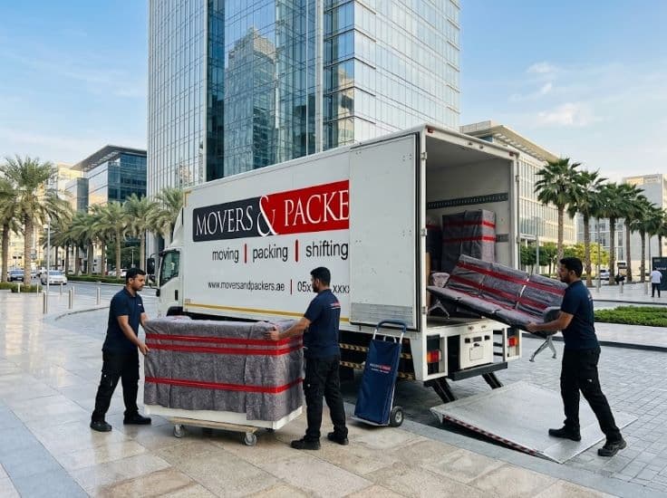 Movers & Packers branded truck being loaded with bank office furniture outside a modern Dubai financial district building