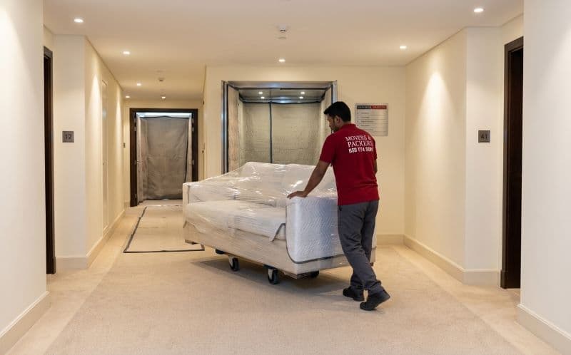 Professional mover carefully transporting a wrapped sofa through a Dubai Marina high-rise corridor toward a padded service lift